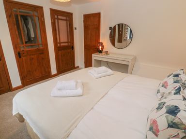 A bedroom with a bed covered in white linen and towels on top wooden doors and a white dressing table with a round mirror and a lamp at Wood Brook Cottage in Crowan