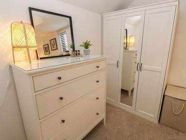 A bedroom with a white chest of drawers with a mirror and lamp next to a white wardrobe with a mirror and a storage basket at Wood Brook Cottage in Crowan