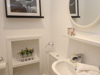 A bathroom sink with a hand towel and soap dispenser a round mirror above and a shelf with a candle a toilet below and flower decorations on a recessed shelf