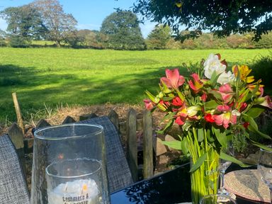 A garden table with a vase of flowers and glass candle holders with chairs and a green field and trees in the background at Wood Brook Cottage in Crowan