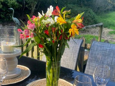 A glass vase with mixed colorful flowers on a black table outdoors with glass candle holder and glasses at Wood Brook Cottage in Crowan