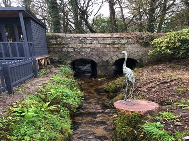 A small stream flowing under a stone bridge next to a dark wooden shed and a metal bird sculpture on a tree stump at Wood Brook Cottage in Crowan