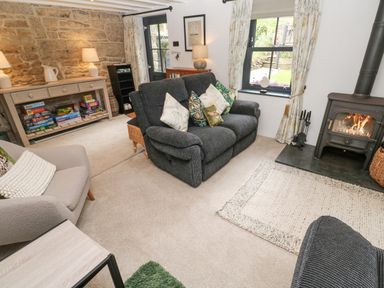 A living room with a sofa and armchair near a wood stove and a console table with board games at Wood Brook Cottage in Crowan