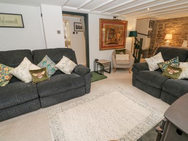 A living room with two dark sofas with decorative pillows a beige armchair side tables framed artwork a stone wall and a rug on the floor at Wood Brook Cottage in Crowan