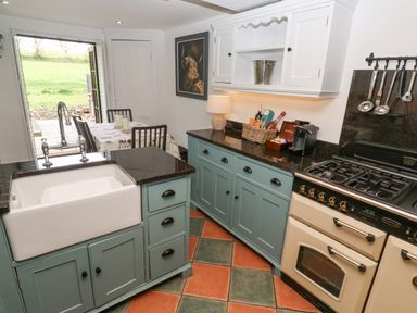 A kitchen with a farmhouse sink stove and dining table near an open door to a garden at Wood Brook Cottage in Crowan