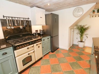 A kitchen with a stove, hanging utensils, refrigerator, potted plant, wall clock, and tiled floor at Wood Brook Cottage Crowan