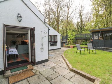 An outdoor patio with an open door leading to a kitchen a grassy area with two chairs and a wooden shed at Wood Brook Cottage in Crowan