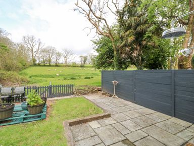 An outdoor patio area with stone tiles and a grassy section next to a black wooden fence and trees with a green field and sheep in the background at Wood Brook Cottage in Crowan