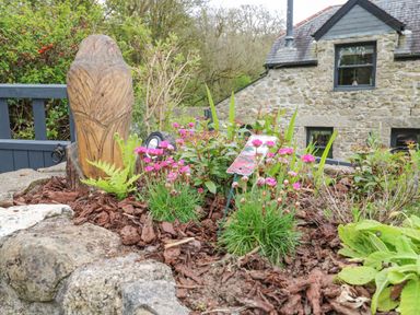 A garden bed with pink flowers a wooden carving and a stone house in the background at Wood Brook Cottage in Crowan