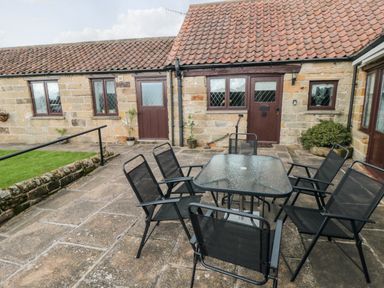 An outdoor patio with a glass table and six black chairs in front of a stone building with brown doors and windows at Beaconsfield Cottage in Burniston near Scalby