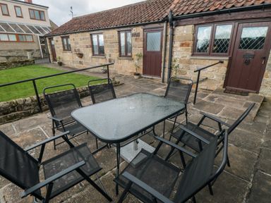A stone patio with a glass table and six black mesh chairs outside a building at Beaconsfield Cottage in Burniston near Scalby
