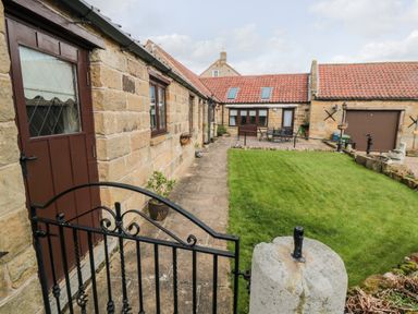 An outdoor courtyard with a grass lawn surrounded by stone buildings with red tile roofs and a black metal gate at Beaconsfield Cottage in Burniston near Scalby