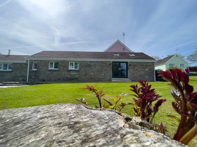 A stone house with windows and a glass door on a lawn with plants growing near a log at The Byre @ Canllefaes in Penparc near Cardigan