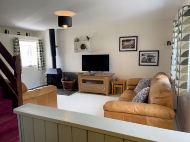 A living room with a tan sofa and armchair a television on a wooden stand a wood-burning stove and patterned curtains at The Shippen @ Canllefaes Penparc near Cardigan