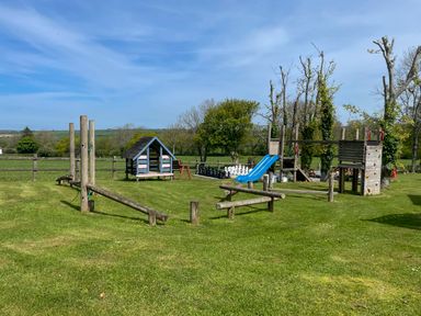 An outdoor playground with wooden climbing structures a blue slide a small playhouse and a large chess set on grass at The Shippen @ Canllefaes in Penparc near Cardigan