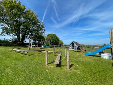 A playground with swings, slides, wooden balancing beams, and a small playhouse on grass at The Shippen @ Canllefaes in Penparc near Cardigan