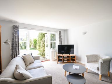 A living room with a sofa an armchair a TV on a stand and double doors leading to a garden at 12 The Gardens in Rousdon