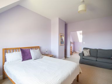 A bedroom with a wooden bed with pillows a bedside table a blue sofa and an open doorway showing a dressing area at 12 The Gardens in Rousdon