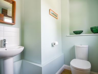 A bathroom with a toilet, sink, wall mirror, toilet paper holder, and decorative bowls on a shelf at 12 The Gardens in Rousdon