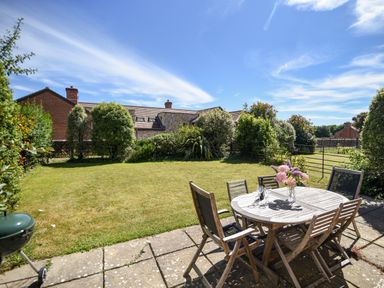 An outdoor patio with wooden table and chairs a vase with flowers on the table and a lawn surrounded by bushes and houses at 12 The Gardens in Rousdon
