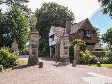 A stone gate entrance with pillars and an iron gate next to a tiled roof house surrounded by trees and bushes at 12 The Gardens in Rousdon