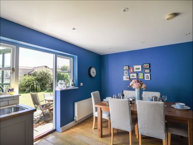 A dining room with a wooden table and white chairs next to a blue wall with picture frames and an open door to a garden at 12 The Gardens in Rousdon