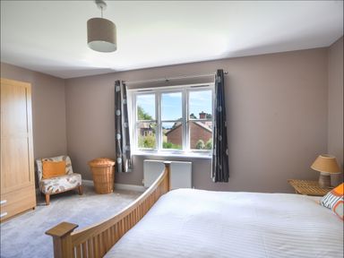 A bedroom with a wooden bed white bedding a chair with an orange pillow a wicker basket a wooden wardrobe and a window with curtains at 12 The Gardens in Rousdon