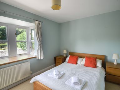 A bedroom with a double bed with white bedding and red cushions two bedside tables with lamps and a window with white curtains at 12 The Gardens in Rousdon