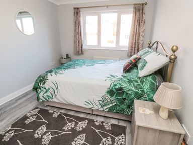 A bedroom with a bed with green leaf patterned bedding a nightstand with a lamp a round mirror and floral curtains at 20 Southmead in Amble