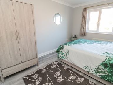 A bedroom with a bed covered in a green and white leaf patterned duvet a wooden wardrobe a round mirror on the wall and a window with curtains at 20 Southmead in Amble