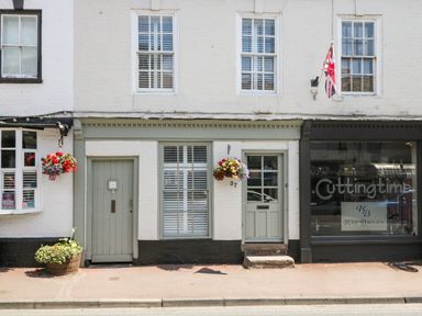 Shopfront with hanging flower baskets and two doors next to a hair salon with a union jack flag above the door at Cuckoo Cottage in Upton Upon Severn