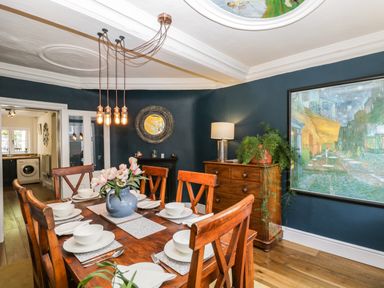 A dining room with a wooden table set for six with white dishes a blue vase with flowers wooden chairs a sideboard with a lamp and plant dark blue walls and a large framed painting at Cuckoo Cottage in Upton Upon Severn