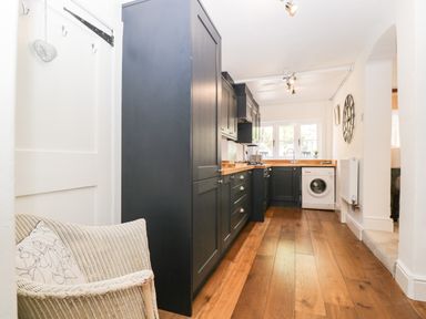 A kitchen with dark cabinets wooden floors a washing machine and a wicker chair with cushions at Cuckoo Cottage in Upton Upon Severn