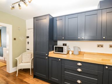 A kitchen with dark cabinets wooden countertop microwave kettle and a white wicker chair at Cuckoo Cottage in Upton Upon Severn