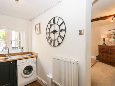 A kitchen area with a washing machine under a wooden counter a large wall clock and a doorway leading to a room with a wooden dresser and lamp at Cuckoo Cottage in Upton Upon Severn