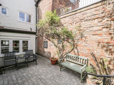 An outdoor patio area with a brick wall two black chairs with a small table between them a green bench and potted plants at Cuckoo Cottage in Upton Upon Severn