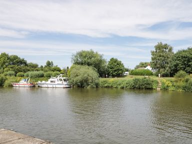 A river with two boats docked near a grassy riverbank with trees and shrubs at Cuckoo Cottage in Upton Upon Severn