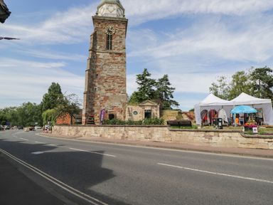 A street view with a tall stone clock tower a small building a white tent with clothes and a blue umbrella on a grassy area
