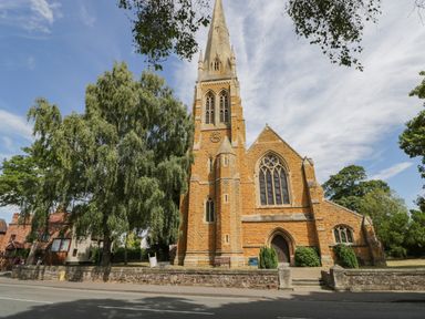 A stone church with a tall spire and arched windows surrounded by trees and a low stone wall at Cuckoo Cottage in Upton Upon Severn