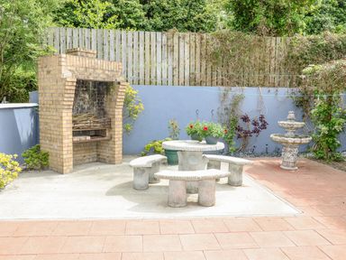An outdoor patio area with a brick barbecue a round stone table with curved benches potted flowers and a stone fountain at 12 Hillview Ludden near Buncrana
