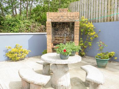 An outdoor patio area with a round stone table and curved stone benches with plants on the table and a brick barbecue in the background at 12 Hillview Ludden near Buncrana