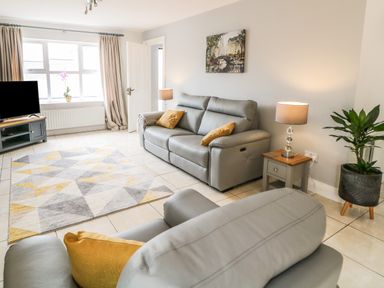 A living room with grey sofas yellow cushions a patterned rug a side table with lamp a plant and a television near a window at 12 Hillview Ludden near Buncrana