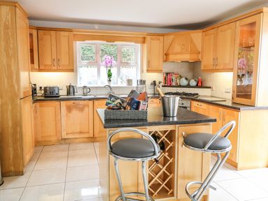 A kitchen with wooden cabinets and black countertops a window with a pink flower a kitchen island with a wine rack two stools and kitchen appliances at 12 Hillview in Ludden near Buncrana