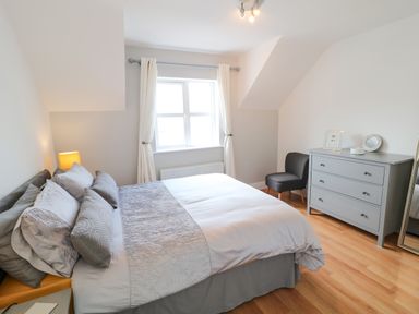A bedroom with a bed, nightstand with lamp, dresser with decorations, chair, and window with curtains at 12 Hillview Ludden near Buncrana