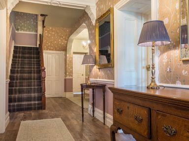 A hallway with wooden furniture table lamps a carpeted staircase and floral wallpaper at Swinside Lodge in Newlands Valley near Keswick
