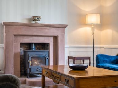 A living room with a wood-burning stove in a stone fireplace a wooden coffee table with a bowl a blue sofa a floor lamp and a small wooden side table at Swinside Lodge in Newlands Valley near Keswick