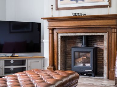 A living room with a wooden fireplace stove a large television on a stand and a brown tufted leather ottoman at Swinside Lodge in Newlands Valley near Keswick