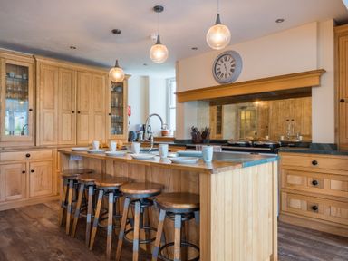 A kitchen with wooden cabinets a wooden island with stools plates and mugs on the island and pendant lights at Swinside Lodge in Newlands Valley near Keswick