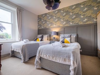 A bedroom with two single beds with gray blankets and pillows a window with curtains and a bedside table with a lamp at Swinside Lodge in Newlands Valley near Keswick