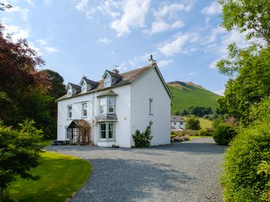 A white house with dormer windows and a gravel driveway surrounded by green trees and a grassy hill in the background at Swinside Lodge in Newlands Valley near Keswick